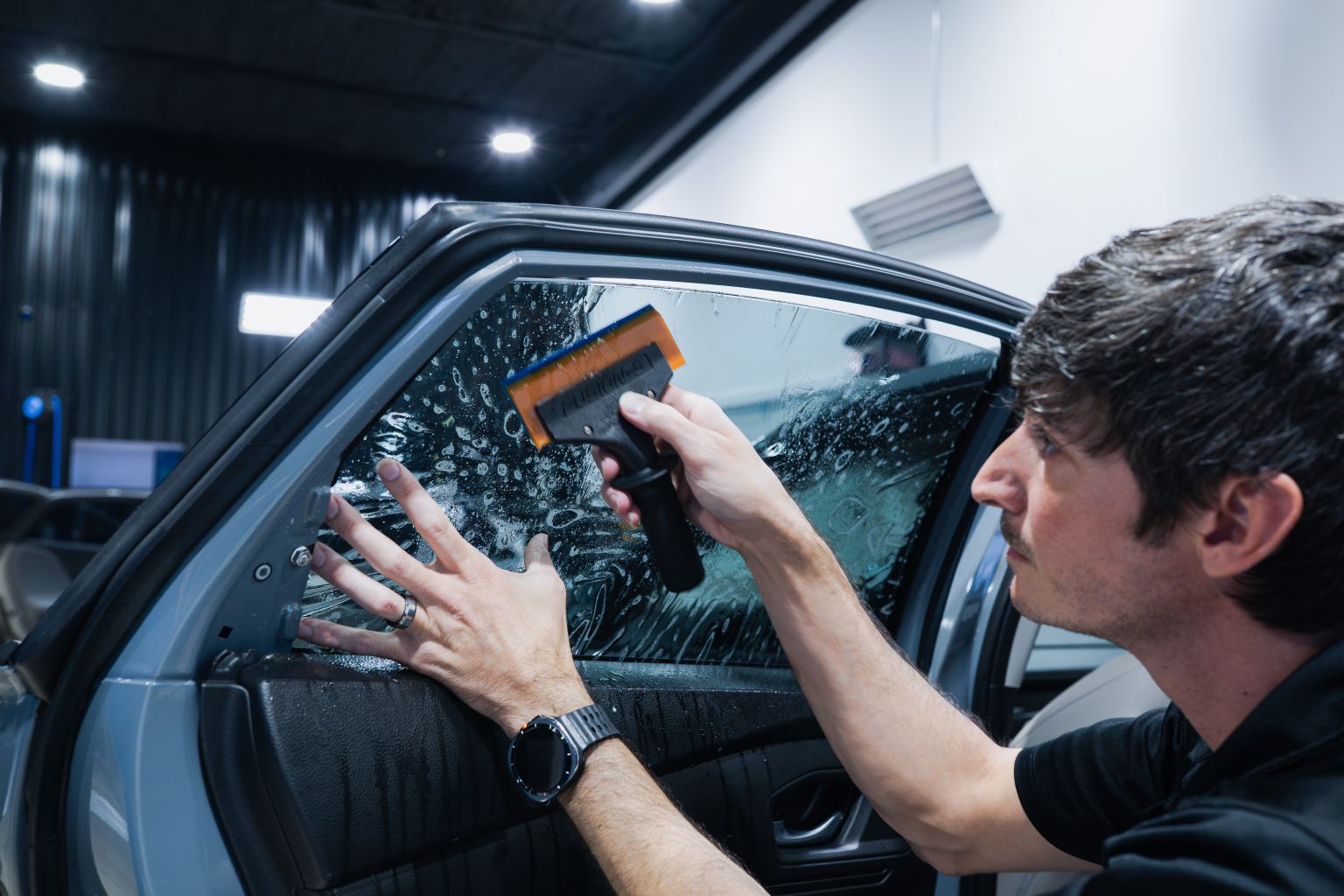 Installer squeegeeing window tint film onto the rear door glass of a gray sedan