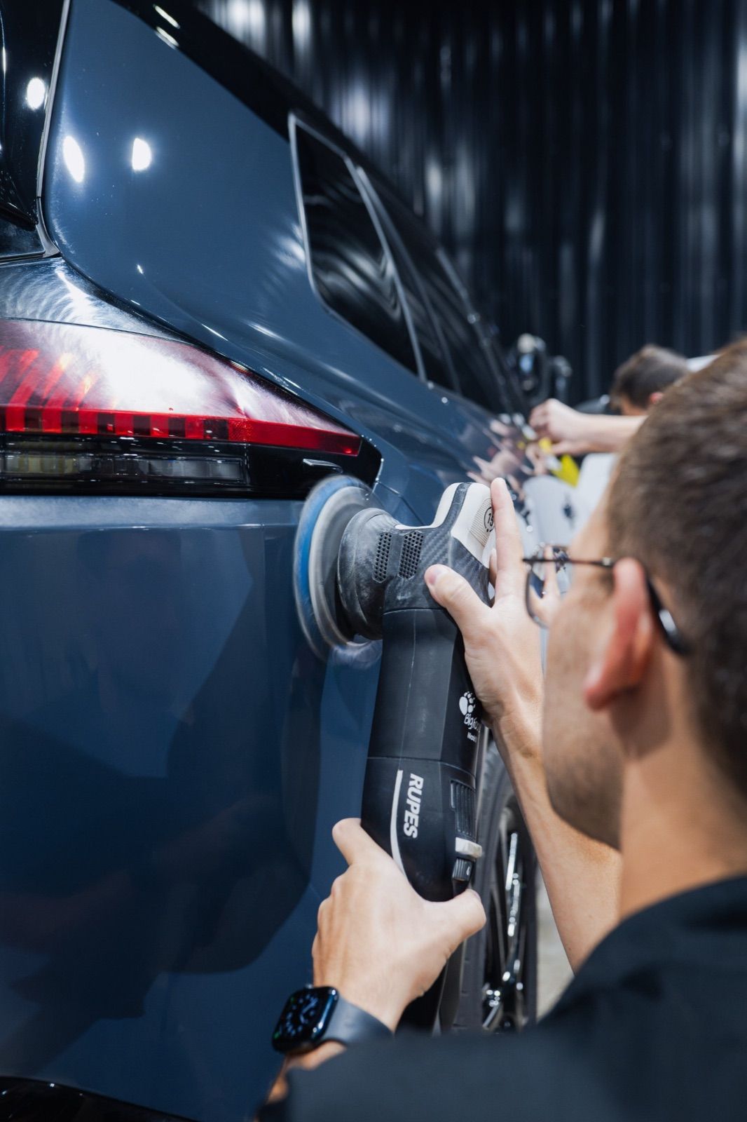 Technician machine-polishing the rear quarter panel of a dark blue SUV with a RUPES polisher