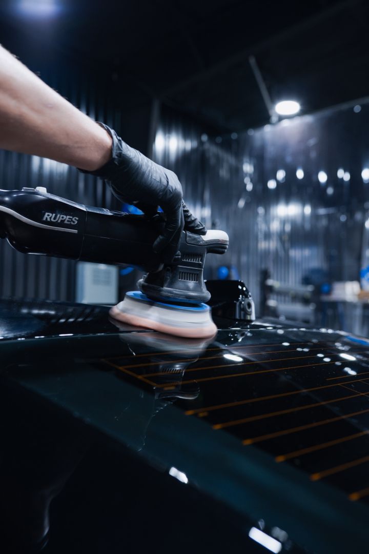 Gloved hand grips a Rupes polishers tool to buff a car hood in a dim workshop with bright overhead lights nearby.