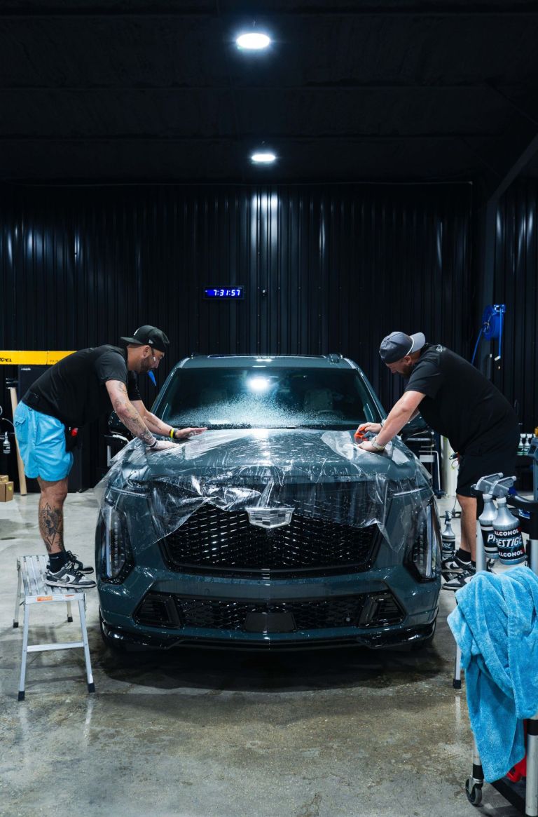 Two Prestige Films installers laying paint protection film across the hood of a gray Cadillac SUV