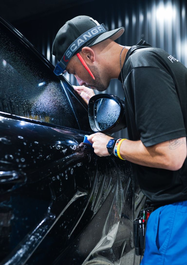 Installer with a headlamp checking paint protection film along the door and mirror of a black car