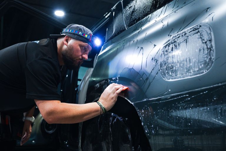 Installer with a headlamp squeegeeing paint protection film on the rear quarter panel of a gray vehicle